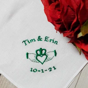 May include: White handkerchief with green embroidered text and a Claddagh design. The text reads "Tim & Erin" above the Claddagh symbol, with the date "10-1-21" below. A bouquet of red roses is in the background.