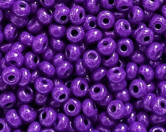 May include: A large collection of small, round, opaque purple glass beads. Each bead features a smooth, glossy finish and a central hole for threading. The beads are uniform in shape and size, creating a dense, vibrant purple texture.