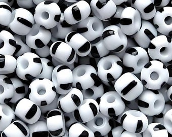 May include: A pile of white beads with black stripe and dot patterns. The beads are round with a hole through the center, and are scattered across the image. These beads are suitable for jewelry making and crafts.
