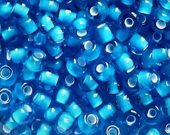 Puede incluir: Una colección de pequeñas cuentas de vidrio azul translúcido de forma redonda. Cada cuenta presenta una superficie lisa y brillante con un orificio central para enhebrar. Las cuentas están densamente agrupadas, creando un campo uniforme de azul.