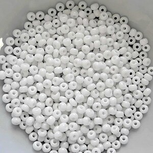 May include: A large quantity of white seed beads, also known as pony beads, in a white bowl. The beads are small and round with a hole in the center.