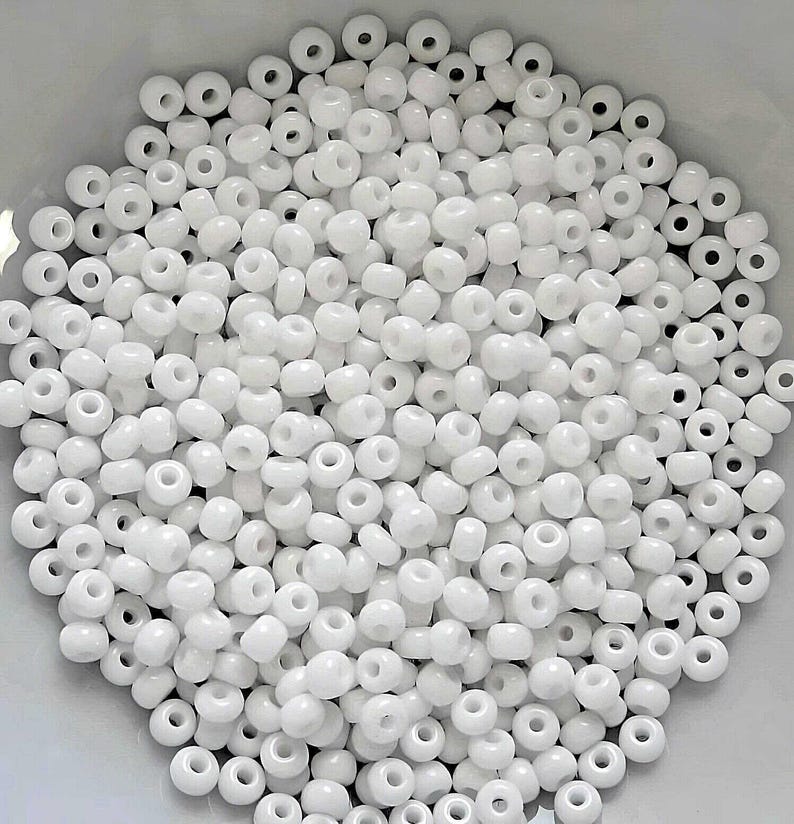 May include: A large quantity of white seed beads, also known as pony beads, in a white bowl. The beads are small and round with a hole in the center.