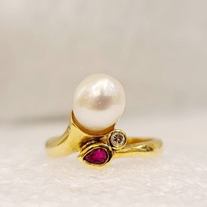 May include: A gold ring with a large white pearl, a small ruby, and a diamond. The ring is designed with a curved band.