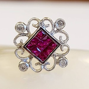 May include: A square ruby gemstone set in a silver filigree design with four smaller diamonds surrounding the ruby.