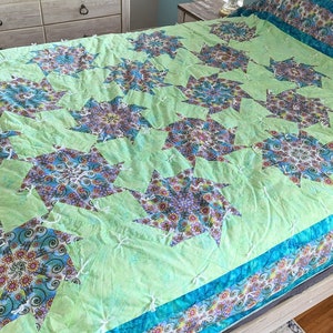 May include: A green quilt with a blue and white floral pattern. The quilt is made up of many smaller squares that are sewn together to create a larger design. The quilt is on a bed with a wooden headboard.