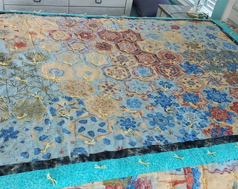 May include: A large, colorful quilt with a patchwork design. The quilt features a variety of patterns, including floral and geometric shapes, in shades of blue, red, and tan. The edges are trimmed with a turquoise border and yellow stitching.