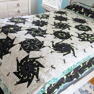 May include: A white and black quilt with a green border. The quilt is made up of a repeating pattern of black and green pinwheels. The quilt is on a bed with a white headboard.