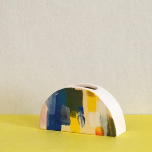 May include: A half-moon shaped ceramic vase with a white exterior and a colorful painted design. The design features brushstrokes of blue, green, yellow, and pink. The vase sits on a yellow surface.