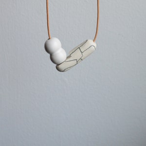 May include: A white ceramic necklace with a geometric pendant and two round beads. The necklace is on a brown cord.