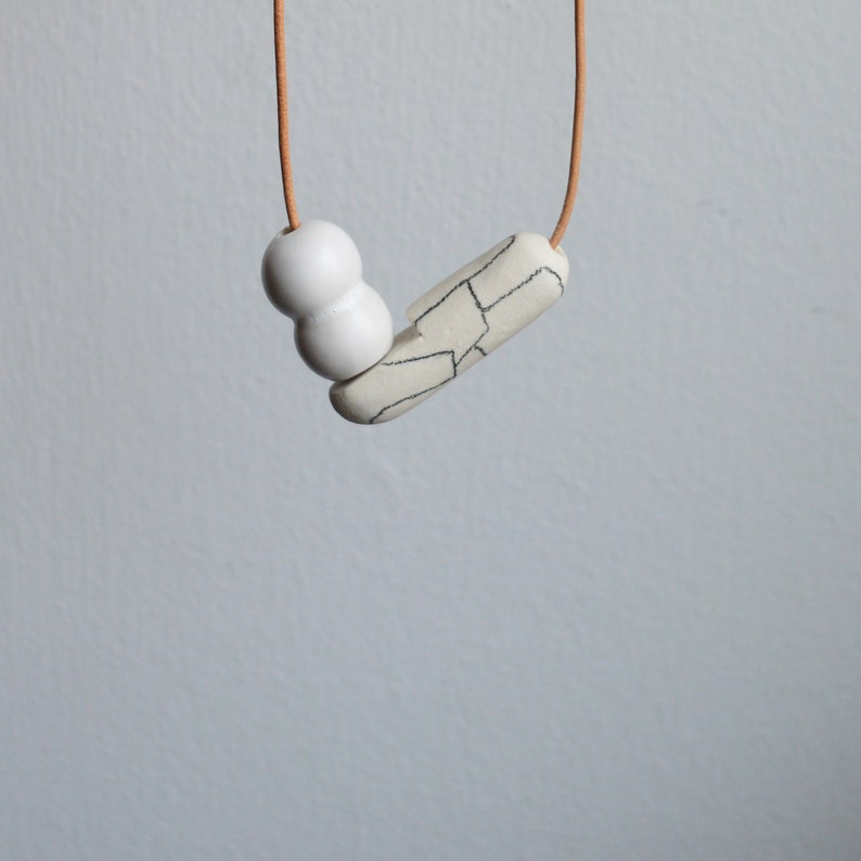 May include: A white ceramic necklace with a geometric pendant and two round beads. The necklace is on a brown cord.