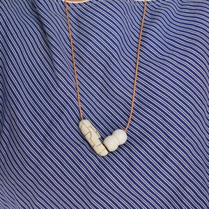 May include: A white ceramic necklace with a geometric pattern and a smooth, round bead. The necklace is on a brown leather cord.