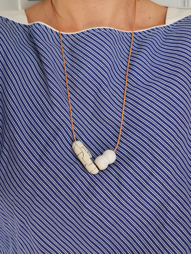 May include: A white ceramic necklace with a geometric pattern and a smooth, round bead. The necklace is on a brown leather cord.