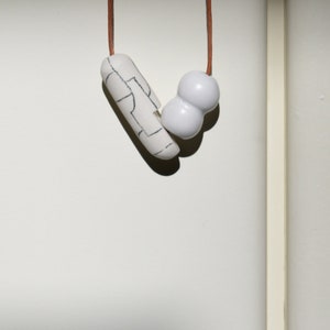 May include: A white ceramic necklace with a long, thin pendant and a round, double-bubble pendant. The necklace is strung on a brown cord.