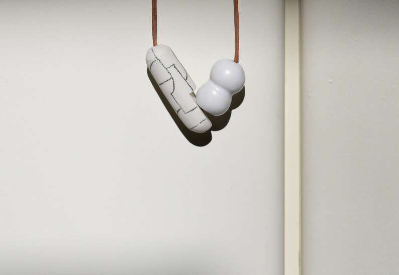 May include: A white ceramic necklace with a long, thin pendant and a round, double-bubble pendant. The necklace is strung on a brown cord.