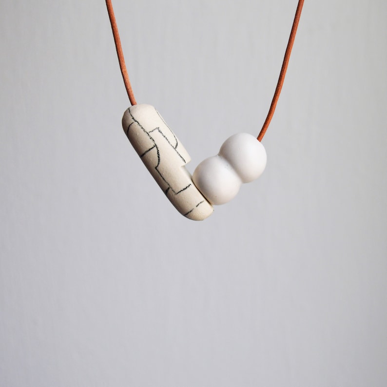 May include: A white ceramic necklace with a brown leather cord. The necklace features a long, rectangular pendant with a black and white geometric pattern and two round beads.