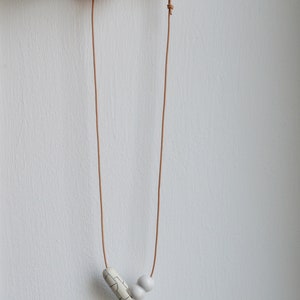 May include: A simple necklace with a brown leather cord and two white ceramic beads. One bead is round and the other is a rectangular shape with a geometric pattern.
