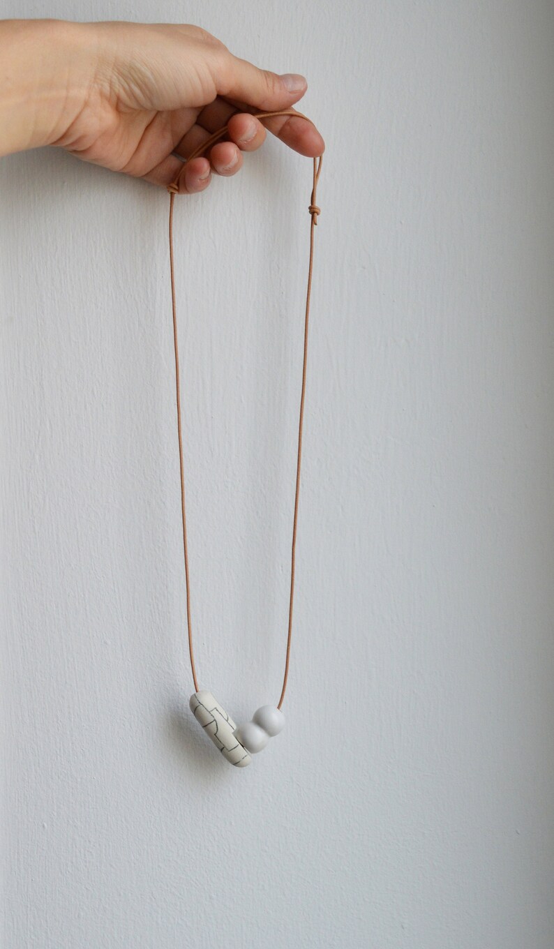 May include: A simple necklace with a brown leather cord and two white ceramic beads. One bead is round and the other is a rectangular shape with a geometric pattern.