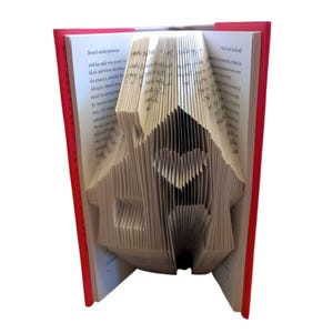 May include: An open book with red covers, its pages folded into the shape of a house. The house design includes a heart-shaped window. The book's pages are white, and the text is visible. The book is a unique decorative item.