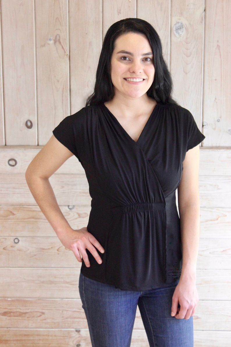 Puede incluir: Una mujer con una blusa negra de manga corta con cuello en V y cintura fruncida. Tambi&eacute;n lleva pantalones vaqueros azules.