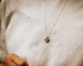 Puede incluir: Un collar de cadena de plata con un pequeño colgante redondo que presenta un diseño texturizado martillado y una pequeña piedra transparente en el centro.