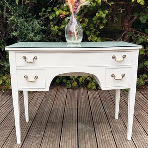 May include: A white vanity desk with a glass top and four drawers featuring gold-toned metal handles. A clear glass vase containing a dried floral arrangement with orange, pink, and purple stems sits on the centre of the desk surface.