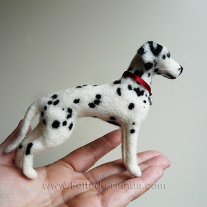 May include: A hand-felted Dalmatian dog figurine with black spots on a white background. The dog is wearing a red collar with a silver charm. The figurine is standing on a person's hand.