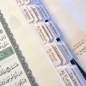 Könnte beinhalten: Nahaufnahme eines weißen und goldenen Koran-Index mit Registerkarten für jedes Kapitel. Die Registerkarten sind mit den Kapiteln in arabischer Schrift und englischer Übersetzung beschriftet. Die Kapitelnamen umfassen "Al Anbiya", "Ash Shu'ara", "Al Baaarah", "Al A'raf", "Al Isra", "Al Haii", "Al Naml", "Ali 'Imran", "Al Kahf", "Al Mu'minun", "An Nisa", "Maryam", "Al Noor", "Ta-Ha", "Al Furqan", "Al 'Ankab" und "Ankaboot".