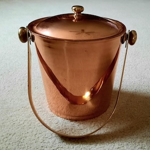 May include: A copper ice bucket with a lid and a handle. The bucket has a shiny, reflective surface and a gold-colored handle. The lid is round with a small knob on top. The bucket is designed to hold ice and keep beverages cold.