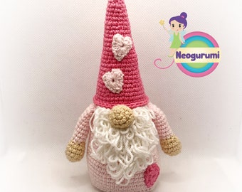 Op de afbeelding: Een roze gehaakte gnome met een witte baard en een roze muts met drie roze harten. De gnome staat op een witte achtergrond.