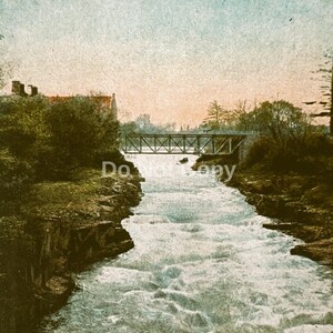May include: A vintage postcard featuring the Union Ave. Bridge over the Passaic River in Little Falls, N.J. The image shows a rushing river flowing between rocky banks toward a metal truss bridge under a pale sky with soft clouds.