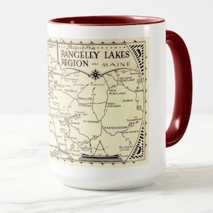 May include: A white ceramic mug with a red rim and handle. The mug features a vintage map of the Rangeley Lakes Region in Maine. The map shows the location of towns and roads in the area. The text "Map of the Rangeley Lakes Region Maine" is printed on the mug.
