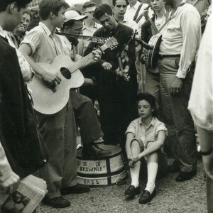 May include: Black and white photograph of a group of people gathered outdoors, with several men playing musical instruments, including guitars and a banjo. A young person sits on the ground, watching the musicians. A barrel with the word "BROWNIE" is in the center.