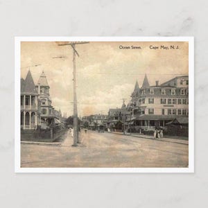 May include: Vintage postcard featuring Ocean Street in Cape May, N.J. The sepia-toned image shows a wide street lined with Victorian-style buildings and a utility pole. The sky is overcast, and a few people are visible.