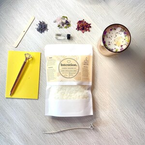 May include: A candle making kit with a white bag of wax flakes, a small bottle of essential oil, dried lavender, rose petals, crystals, a wooden stirring stick, a gold pen, a yellow notepad, a wick, and a wick sticker. The bag has a label that says "Intention. Candle Making Kit Scent: Vanilla Coconut Amber Hand Crafted"