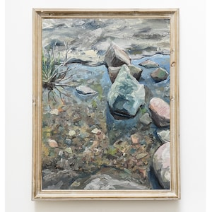 Puede incluir: Pintura al óleo de un arroyo con rocas y agua. La obra de arte presenta una variedad de tamaños y colores de rocas, con un cielo azul claro y gris. La pintura está enmarcada en un marco de madera de color claro.