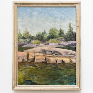 Puede incluir: Pintura al óleo de un paisaje con cielo azul claro, árboles verdes y una costa rocosa. La pintura está en un marco de madera con un acabado marrón claro. El agua es de color verde oscuro.