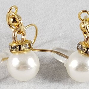 May include: A pair of pearl earrings with gold-colored accents. Each earring features a white, round pearl, a gold-colored decorative cap with clear rhinestones, and a gold-colored hook. The earrings are designed for pierced ears.