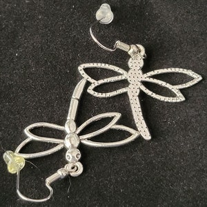 May include: A pair of silver dragonfly earrings with a textured design. The earrings have a hook closure.