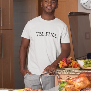 K&ouml;nnte beinhalten: Ein Mann tr&auml;gt ein wei&szlig;es T-Shirt mit dem Aufdruck "I'M FULL". Er steht in einer K&uuml;che mit einem Tisch voller Essen, darunter ein gebratener Truthahn, Kartoffeln und Brot.