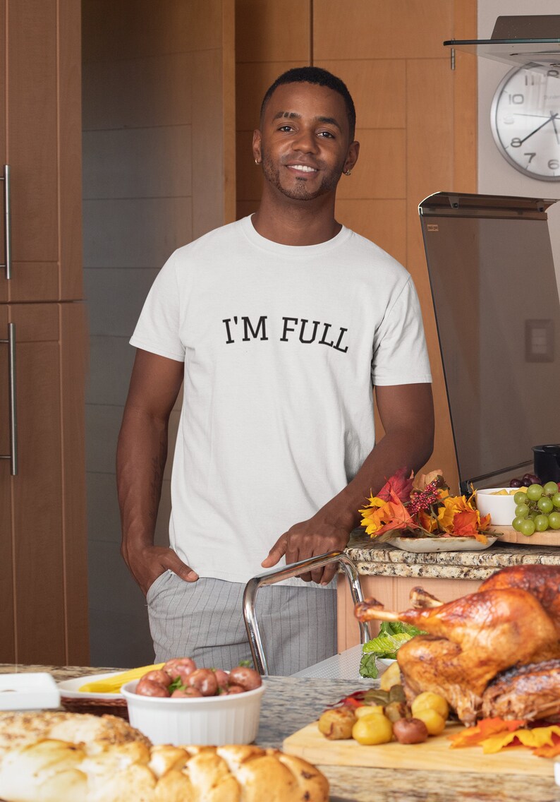 K&ouml;nnte beinhalten: Ein Mann tr&auml;gt ein wei&szlig;es T-Shirt mit dem Aufdruck "I'M FULL". Er steht in einer K&uuml;che mit einem Tisch voller Essen, darunter ein gebratener Truthahn, Kartoffeln und Brot.