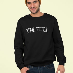 K&ouml;nnte beinhalten: Ein schwarzer Pullover mit dem Text "I'M FULL" in wei&szlig;en Buchstaben. Das Model tr&auml;gt eine braune M&uuml;tze mit Bommel.