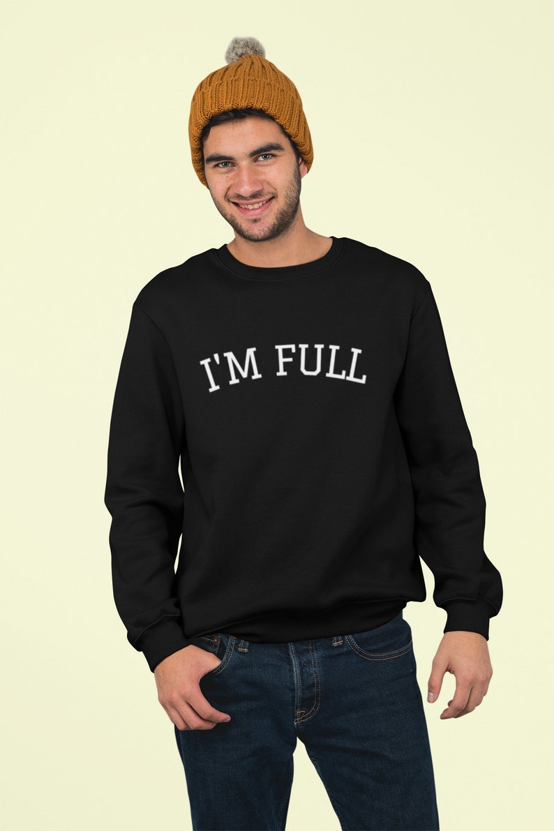 K&ouml;nnte beinhalten: Ein schwarzer Pullover mit dem Text "I'M FULL" in wei&szlig;en Buchstaben. Das Model tr&auml;gt eine braune M&uuml;tze mit Bommel.