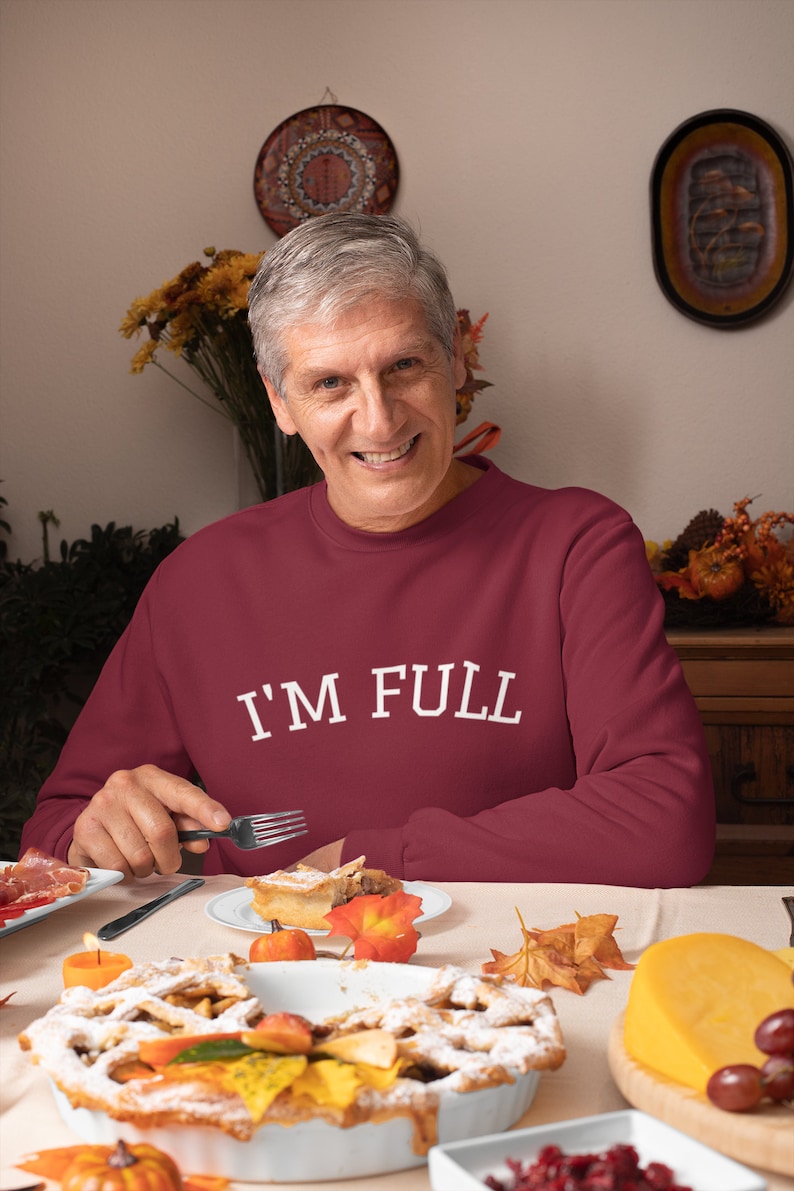 K&ouml;nnte beinhalten: Ein Mann tr&auml;gt ein weinrotes Sweatshirt mit dem Text "I'M FULL" in wei&szlig;en Buchstaben und sitzt an einem Tisch mit einem St&uuml;ck Apfelkuchen auf einem Teller vor ihm. Auf dem Tisch steht ein ganzer Apfelkuchen sowie andere Speisen wie Trauben, K&auml;se und ein K&uuml;rbis.