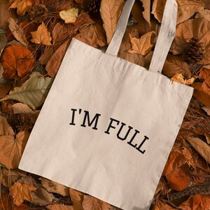 K&ouml;nnte beinhalten: Eine wei&szlig;e Stofftasche mit schwarzem Text, der "I'M FULL" auf einem Bett aus braunen Herbstbl&auml;ttern lautet.