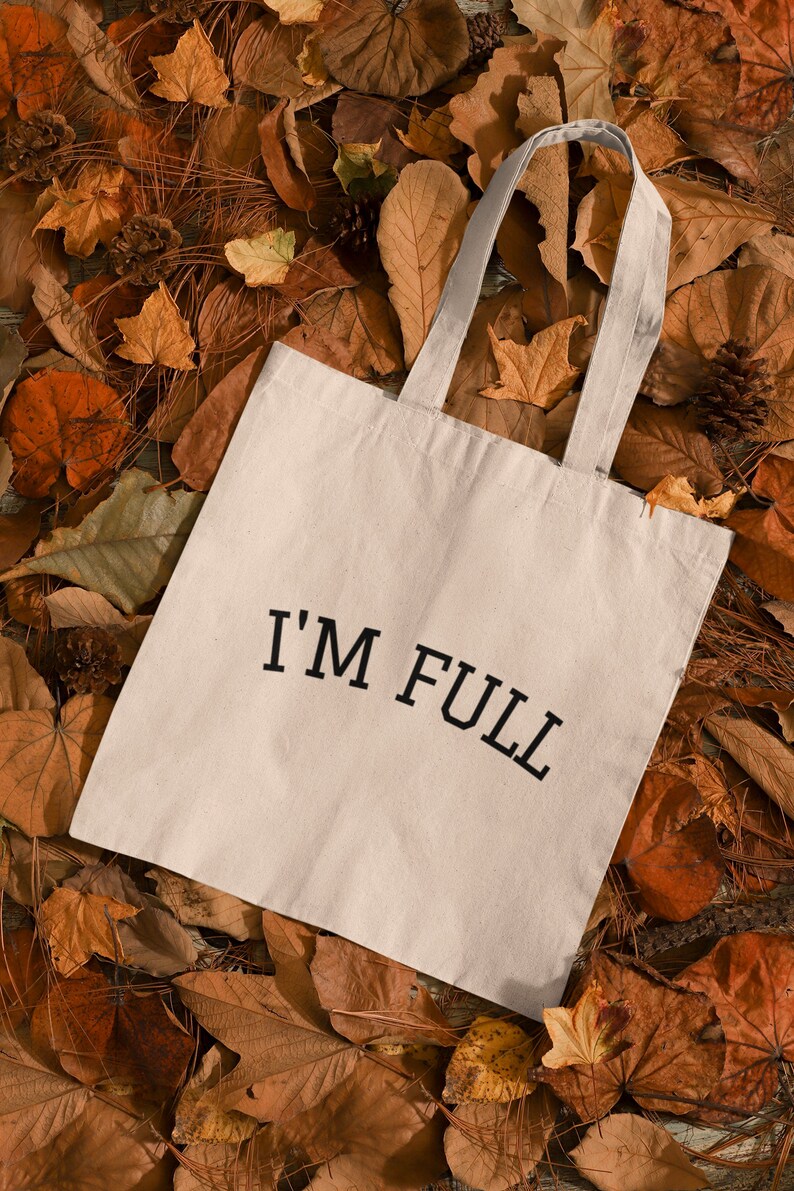 K&ouml;nnte beinhalten: Eine wei&szlig;e Stofftasche mit schwarzem Text, der "I'M FULL" auf einem Bett aus braunen Herbstbl&auml;ttern lautet.