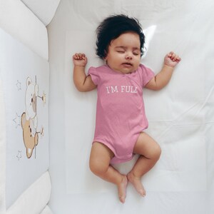 K&ouml;nnte beinhalten: Ein rosa Baby-Strampler mit dem Text "I'M FULL" in wei&szlig;en Buchstaben. Der Strampler liegt auf einem wei&szlig;en Babybettlaken.