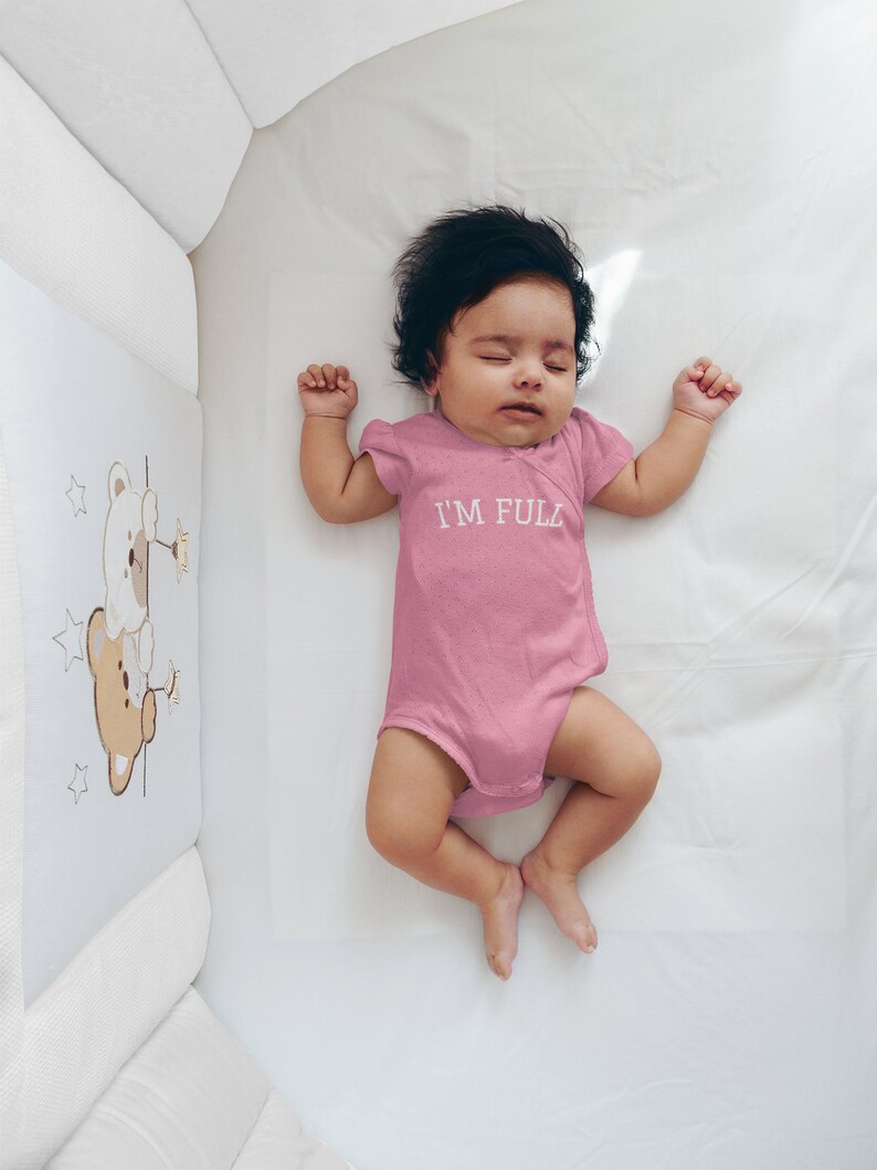 K&ouml;nnte beinhalten: Ein rosa Baby-Strampler mit dem Text "I'M FULL" in wei&szlig;en Buchstaben. Der Strampler liegt auf einem wei&szlig;en Babybettlaken.