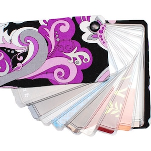 May include: A fabric-covered card holder featuring a black, purple, and white paisley-inspired floral pattern. The accessory includes multiple clear plastic sleeves fanned out to display various cards, secured by a single metal rivet at the top corner.