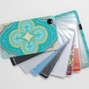 May include: A fan-shaped swatch book with a teal, yellow, and gray patterned cover. The book contains multiple color and pattern samples, including black, gray, blue, and red designs. A black button secures the book.