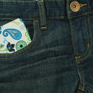 May include: A dark blue denim fabric with yellow stitching, a metal button, and a small pocket containing a white rectangular object featuring a blue and green paisley pattern with floral motifs and a small black circular accent on the left side.